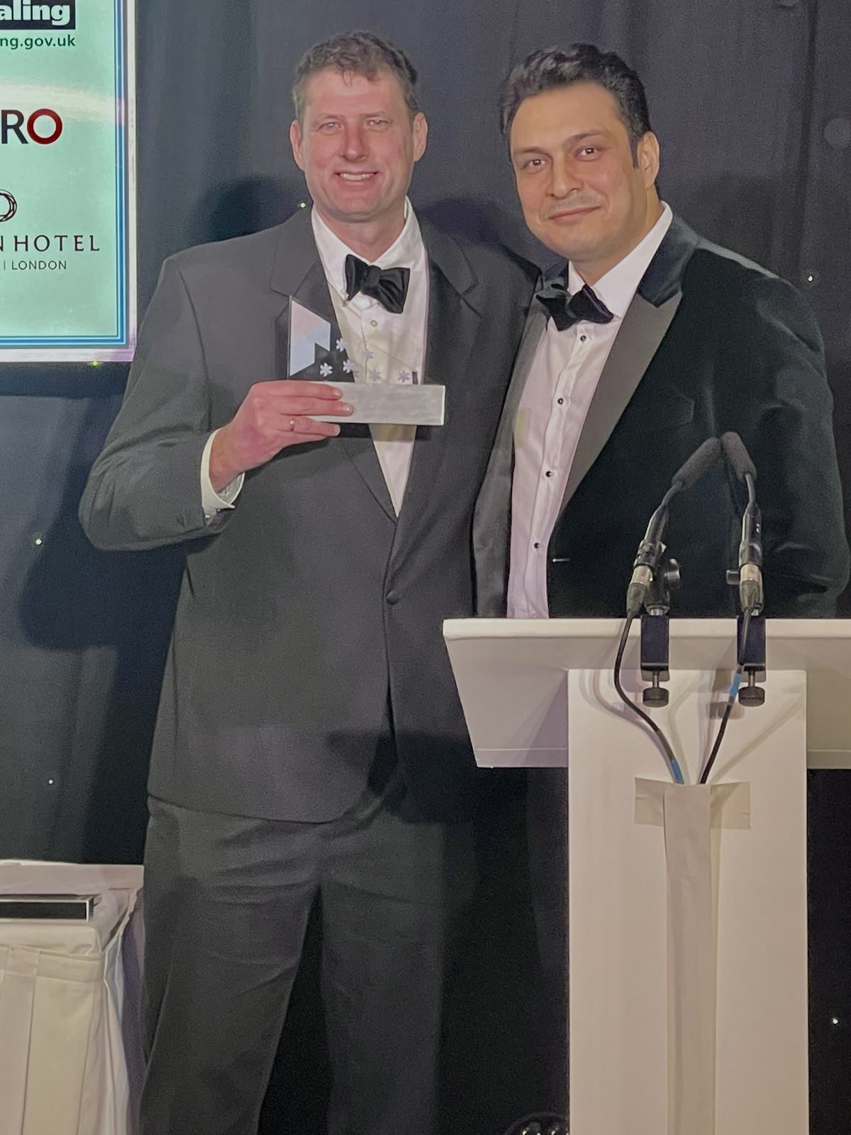 Two men in tuxedos behind a podium holding an award at the West London Business Awards
