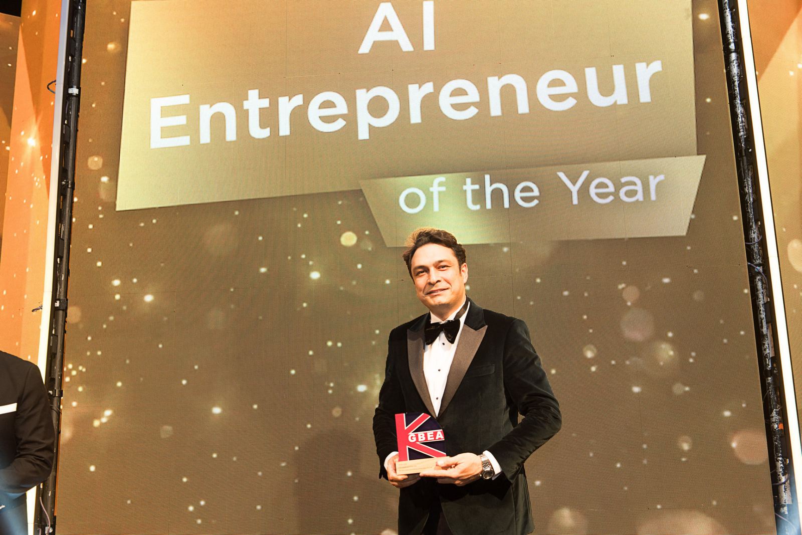 Nima Eskandari holding a GBEA award in front of a screen reading AI Entrepreneur of the Year